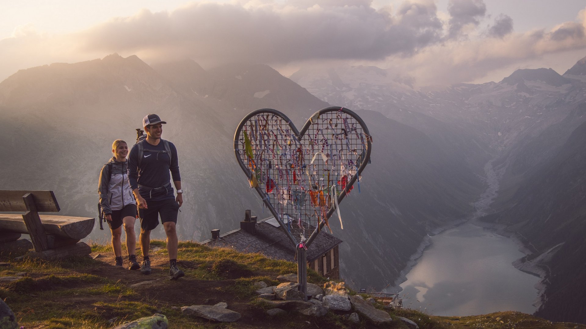 Wanderung bei Sonnenuntergang Zwei Wanderer bei Sonnenuntergang neben einem herzförmigen Gitter mit bunten Bändern, mit Bergsee und Bergen im Hintergrund.