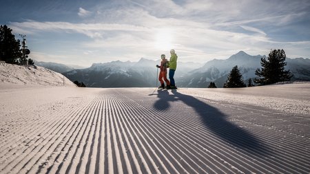 Skifahrer auf präparierter Piste Zwei Skifahrer stehen auf einer frisch präparierten Piste bei Sonnenaufgang und genießen die Aussicht auf die Berge.