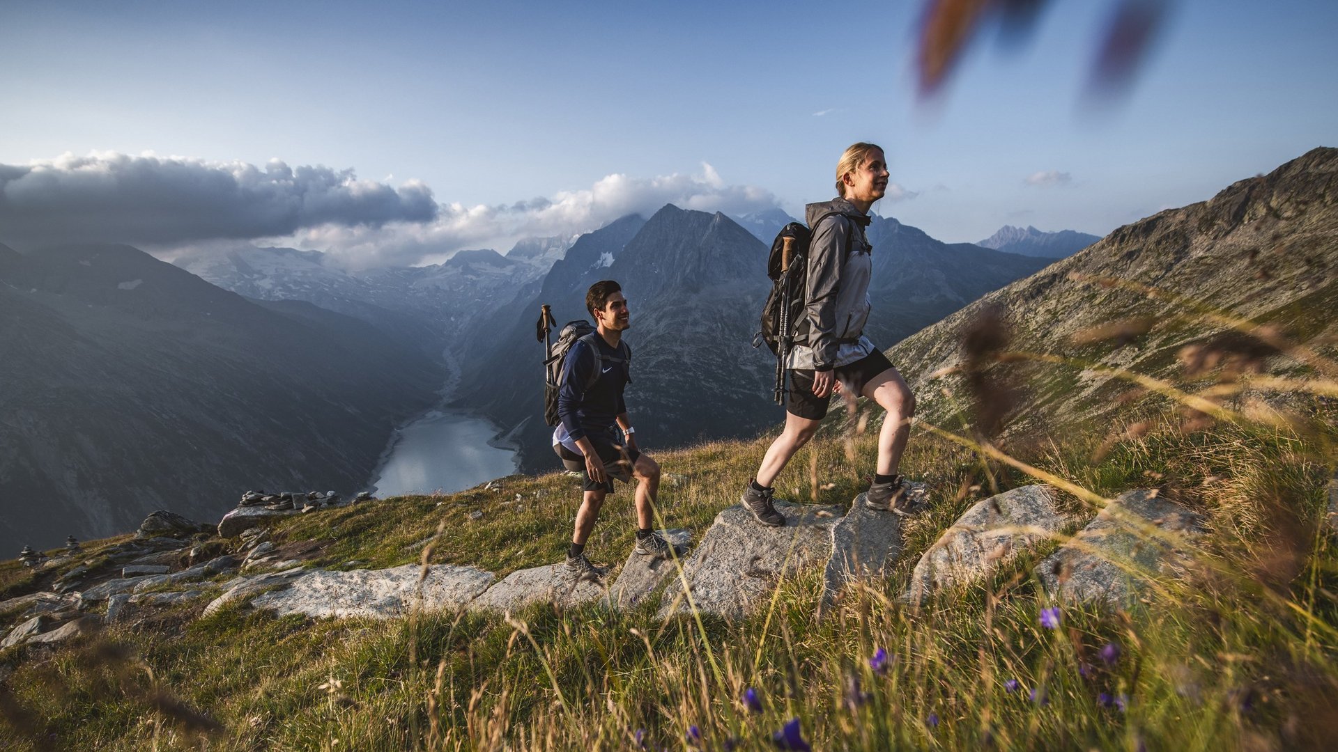 Zwei Wanderer steigen einen Bergpfad hinauf mit einem malerischen Bergsee und Berggipfeln im Hintergrund.