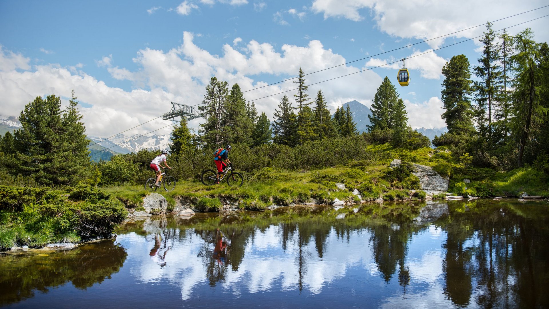 Mountainbiken in den Bergen Zwei Mountainbiker fahren entlang eines Waldpfads neben einem See, während eine Seilbahn über ihnen fährt.