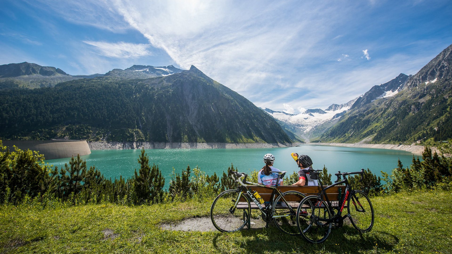 Radpause am Bergsee Zwei Radfahrer sitzen auf einer Bank mit Blick auf einen malerischen Bergsee, umgeben von Bergen und Gletschern.