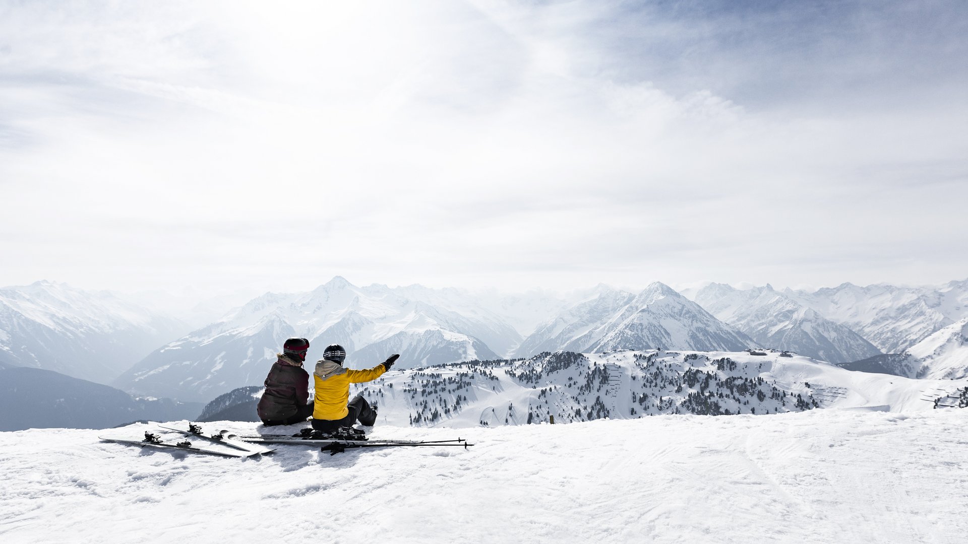 Skifahrer genießen Bergblick Zwei Skifahrer sitzen im Schnee und blicken auf die weitläufige Berglandschaft unter ihnen.