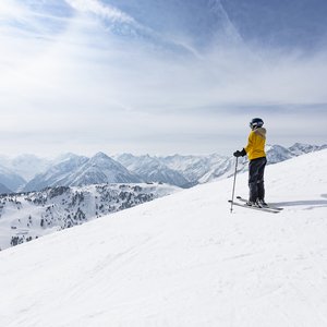 Skifahrer auf Berggipfel Skifahrer in gelber Jacke steht auf einem schneebedeckten Hang und blickt auf die umliegenden verschneiten Berge.