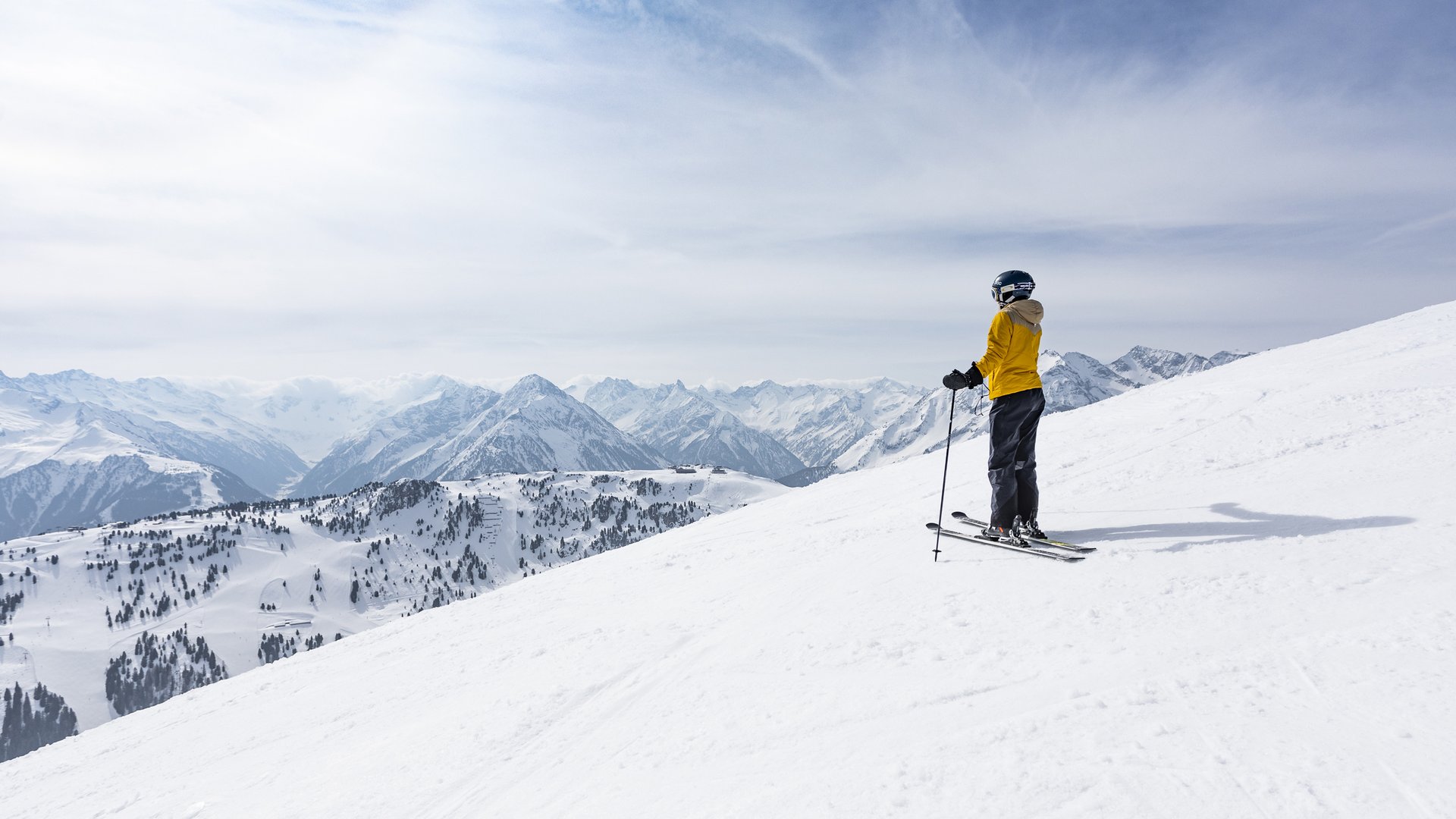 Skifahrer in gelber Jacke steht auf einem schneebedeckten Hang und blickt auf die umliegenden verschneiten Berge.